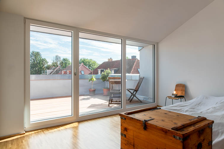 Innenraumperspektive des Schlafzimmers im Dachgeschoss. Blick durch die bodentiefen Fenster auf die Dachterrasse